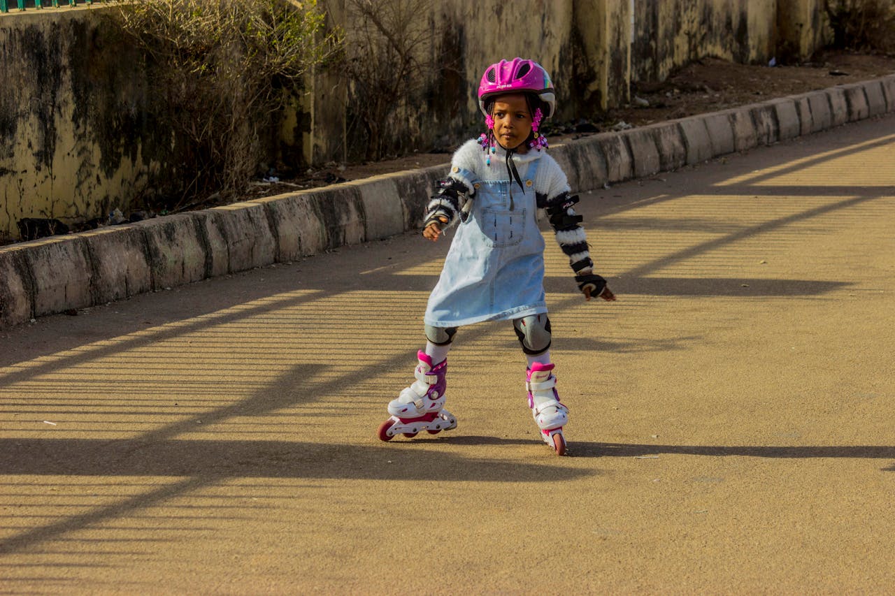 A young girl in protective gear roller skating on a sunny day, wearing a pink helmet and blue dress.